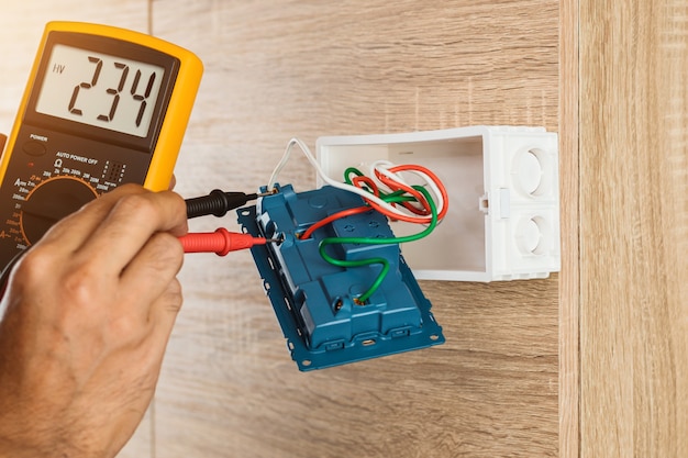 Electrician using a digital meter to measure the voltage at a wall socket on a wooden wall.