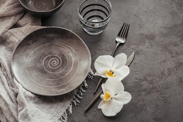 Elegance table setting with knitted grey napkin, cutlery, ceramic plates, glasses and white orchid flowers on dark table. Holiday modern table decoration. Romantic dinner.