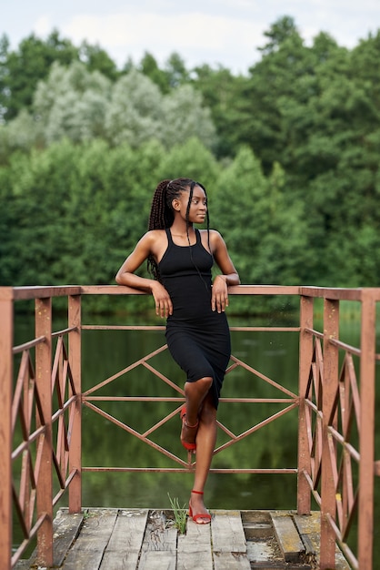 elegant afro woman in fashion dress stands near a blue lake in sunny summer day