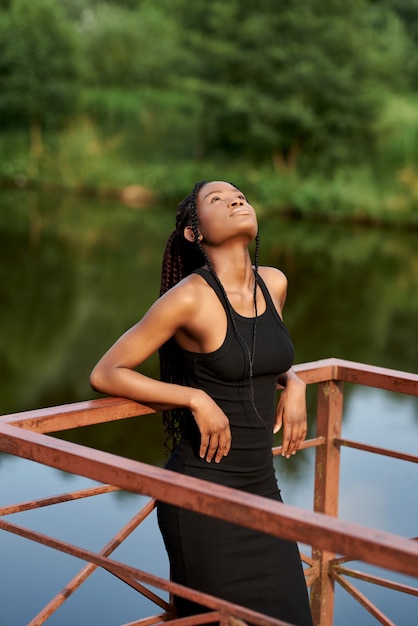 elegant afro woman in fashion dress stands near a blue lake in sunny summer day