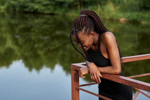 elegant afro woman in fashion dress stands near a blue lake in sunny summer day