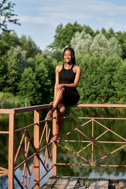 elegant afro woman in fashion dress stands near a blue lake in sunny summer day