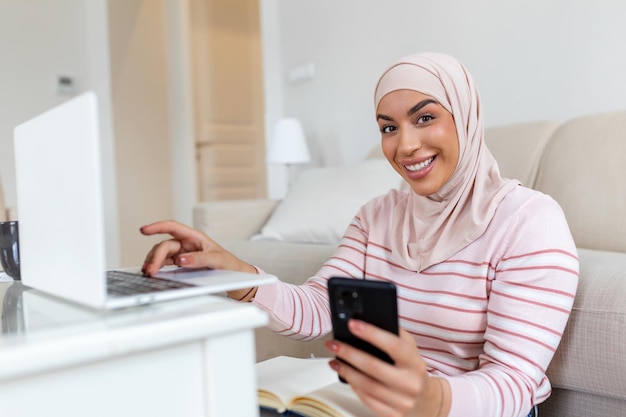 Elegant attractive Muslim woman using mobilephone and laptop searching online shopping information in living room at home Portrait of happy woman purchasing product via online shopping