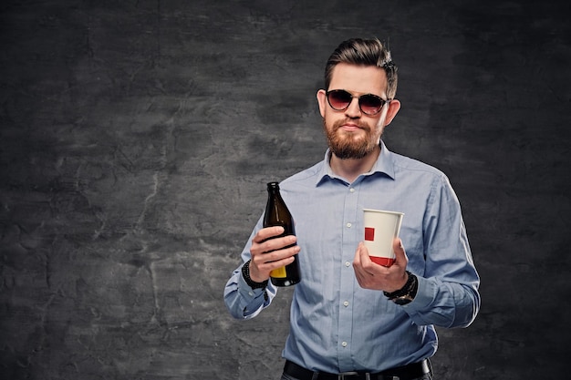 The elegant bearded hipster male holds a paper coffee cup and craft bottled beer.