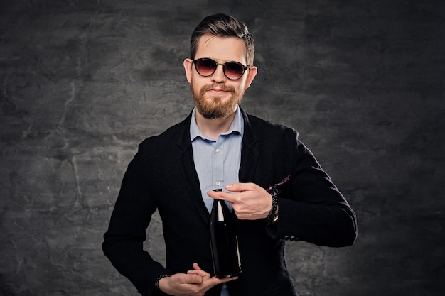 An elegant bearded male dressed in a cotton velvet jacket and sunglasses presenting craft bottled beer.