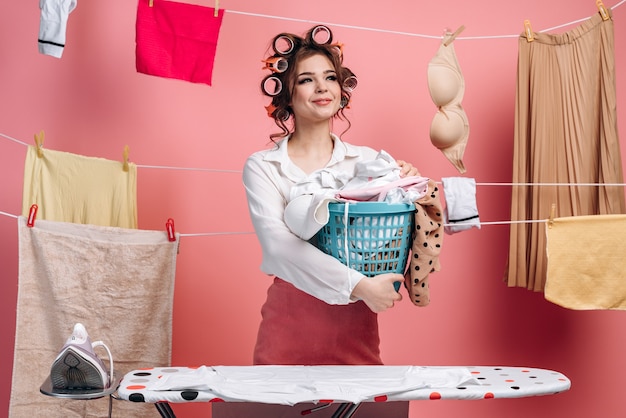 An elegant, beautiful housewife with a curler on her head holds a basket of clothes, behind her clothes hang on a string