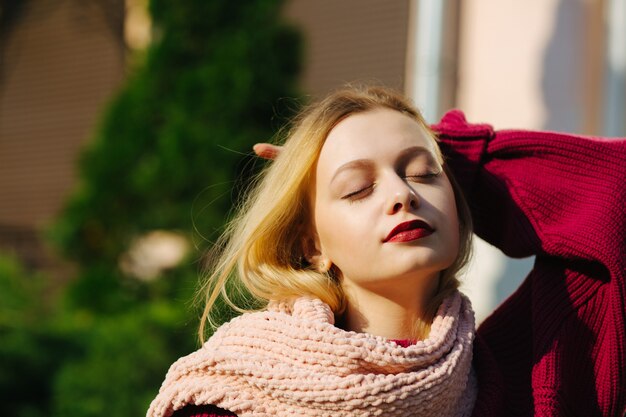 Elegant blonde woman with red lips wearing pink knitted scarf posing in rays of sun. Space for text