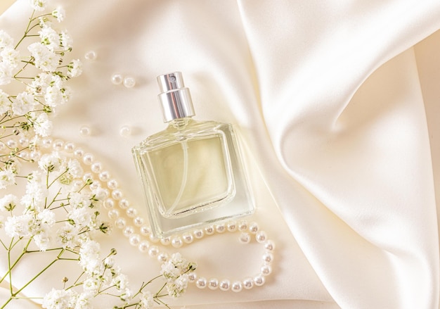 An elegant bottle of women's perfume on ivorycolored atlas fabric with white gypsophila and pearls sensual smell top view