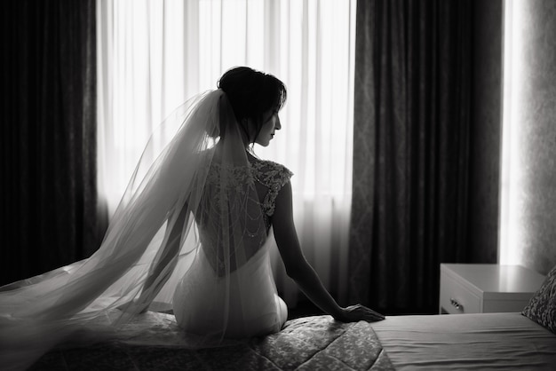 Elegant bride in a wedding dress sits on a bed in a hotel in front of window. Wedding morning of the bride.