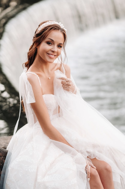 An elegant bride in a white dress, gloves and bare feet is sitting near a waterfall in the Park enjoying nature.A model in a wedding dress and gloves at a nature Park.Belarus