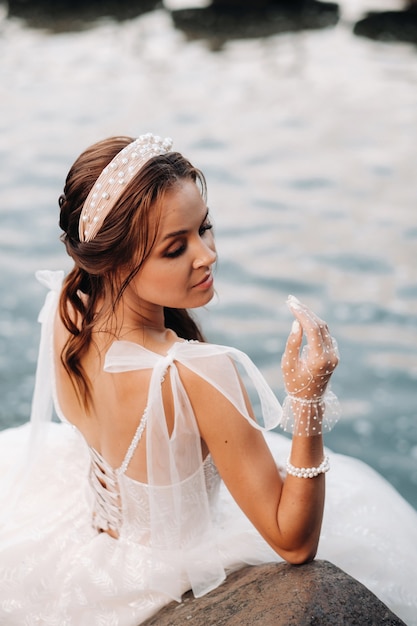 An elegant bride in a white dress, gloves and bare feet is sitting near a waterfall in the Park enjoying nature.A model in a wedding dress and gloves at a nature Park.Belarus