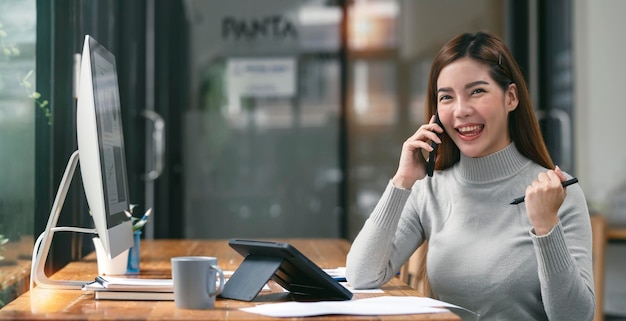 Elegant businesswoman sitting in office using mobile phone and digital tablet Excited asian businesswoman raising hands to congratulate while working on computer in office room