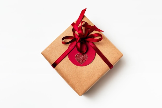 Elegant design of present gift box wrapped in brown craft paper with red ribbon and red tag, top view on white background