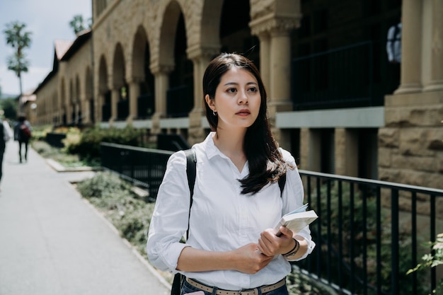 elegant female exchange student holding school book going to course in university. hard working college girl walking outdoor in stanford. serious young woman study abroad in america alone.