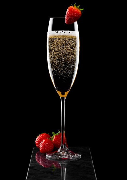 Elegant glass of yellow champagne with strawberry on top and fresh berries on black marble board on black