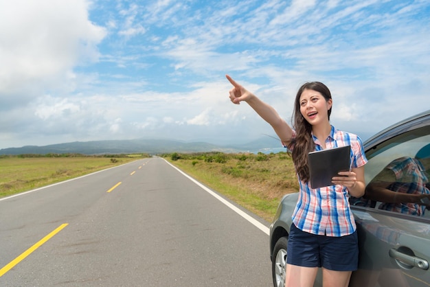 elegant happy female student with car on asphalt road holding digital tablet pad computer searching for local travel information and making pointing gestures sharing beautiful scenery.