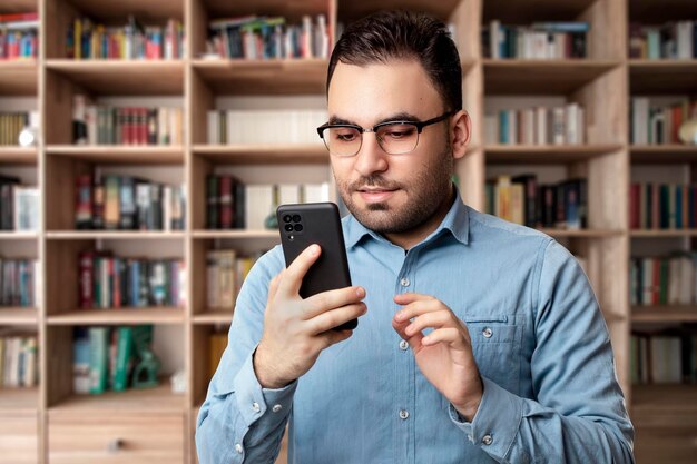 An elegant man standing in the library holding his mobile phone and looking at him wearing glasses.