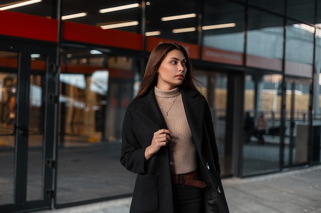 Elegant pretty young brunette woman in a fashionable black coat in a knitted beige shirt stands near a modern glass building on the street. Trendy girl fashion model in stylish clothes in the city.