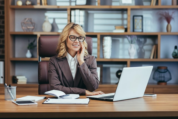 Elegant senior businesswoman with glasses smiling while working on a laptop in a modern