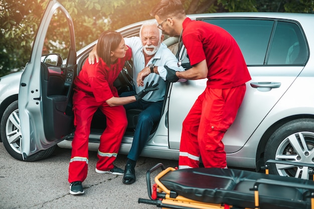 Elegant senior man with heart attack symptoms sitting in the car, emergency medical service workers trying to help him.