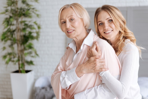 Elegant senior woman covering the hand of her daughter with her own one while the daughter embracing her from behind