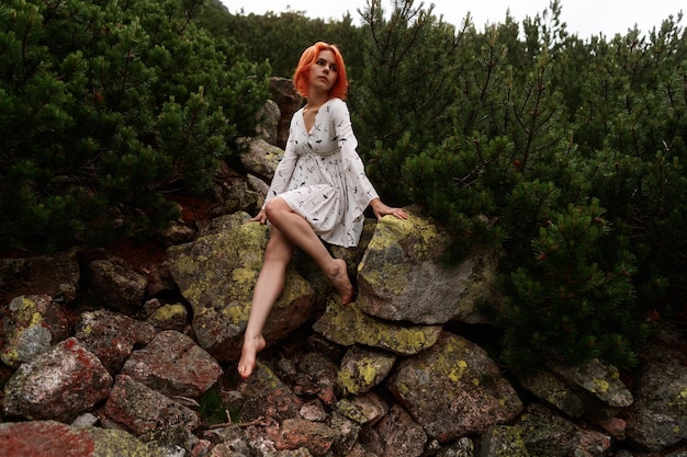 Photo elegant sexy lady with red hair in a white dress sits on the rock among spruce in the mountains