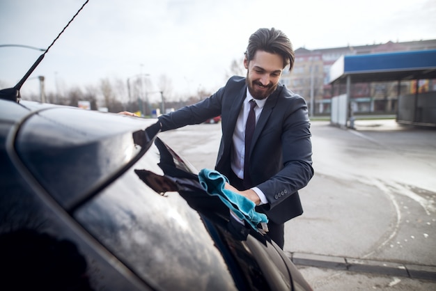 Elegant stylish cheerful young bearded man in a suit cleaning back window of the car with a blue microfiber cloth.