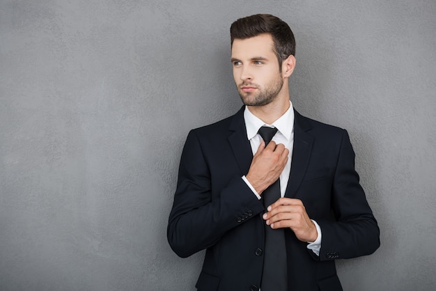 Elegant and successful. Confident young businessman adjusting his necktie and looking away while standing against grey background