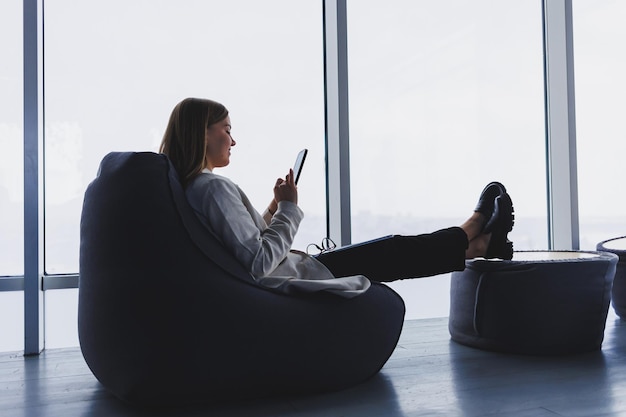 Elegant woman analyzing charts on gadgets and concentrating on phone screen while sitting by full wall window in modern office