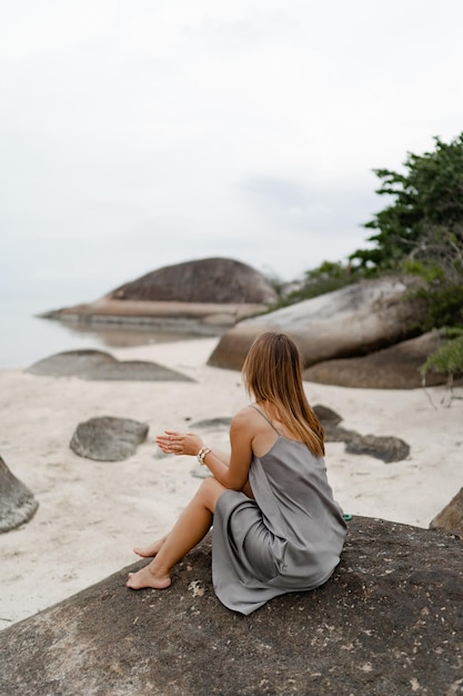 Elegant woman in grey silk dress posing on lonely beach in cloudy weather View from back