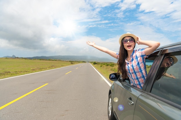 elegant woman wearing hat and sunglasses happily looking out car window open arm enjoying sunshine holiday on asphalt road background.