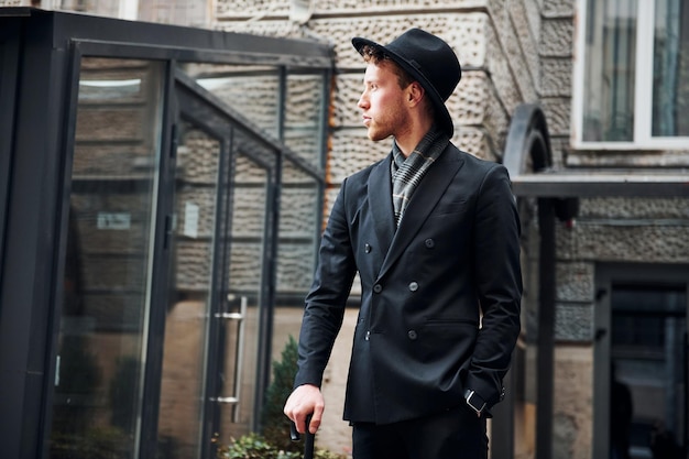 Elegant young man in black hat and clothes with umbrella in hands outdoors in the city