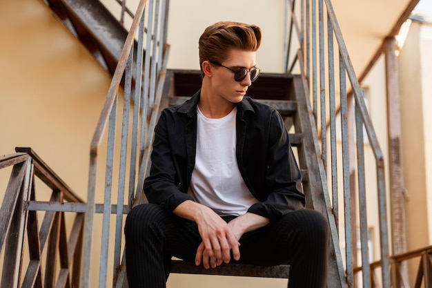 Elegant young man hipster with a hairstyle in a classic black shirt in a white tshirt in stylish pants in sunglasses