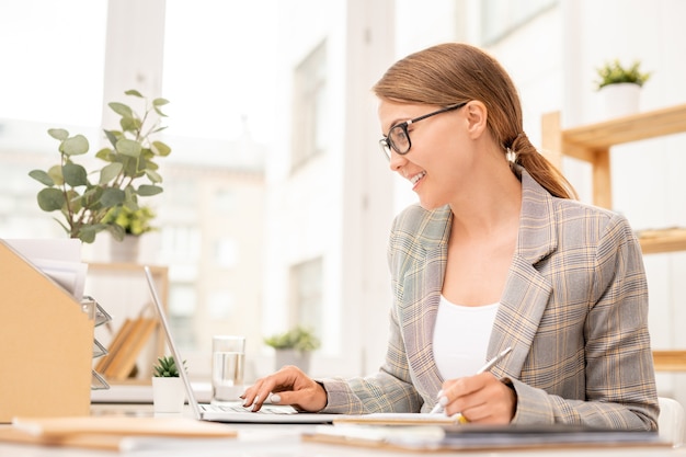 Elegant young office manager making working notes while looking through online data in front of laptop