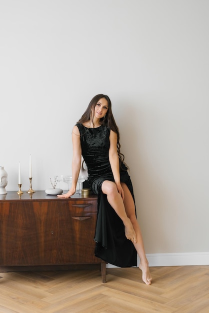 Elegant young woman in a black dress smiles and looks at the camera sitting on a wooden chest of drawers in the living room