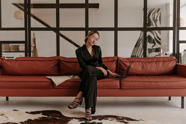 Elegant young woman in black jumpsuit looks into camera and sits on soft brown sofa in living room