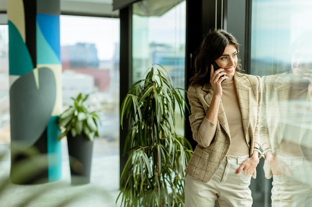 Elegant young woman using mobile phone and contemplates the cityscape through a modern office window