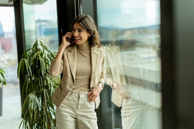 Elegant young woman using mobile phone and contemplates the cityscape through a modern office window