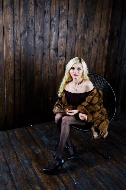 Elegante blonde girl wear on fur coat sitting on chair at studio against wooden background.