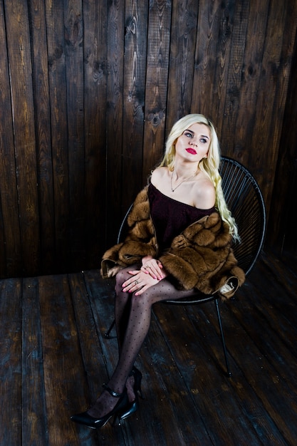 Elegante blonde girl wear on fur coat sitting on chair at studio against wooden background.