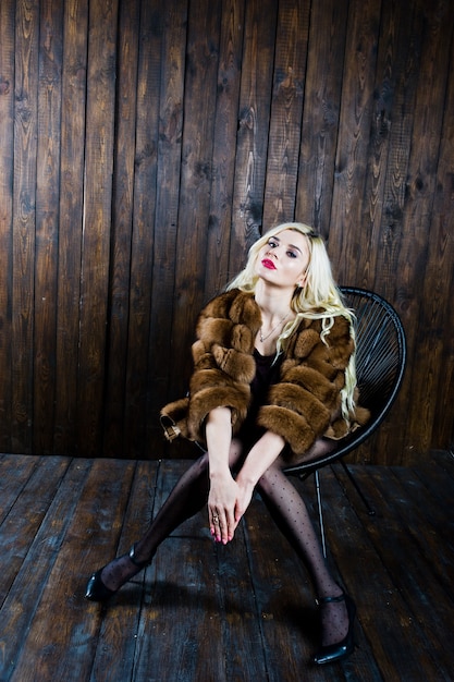 Elegante blonde girl wear on fur coat sitting on chair at studio against wooden background.