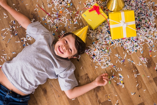 Elevated view of a happy boy lying on hardwood floor with confetti; gift boxes and party hat