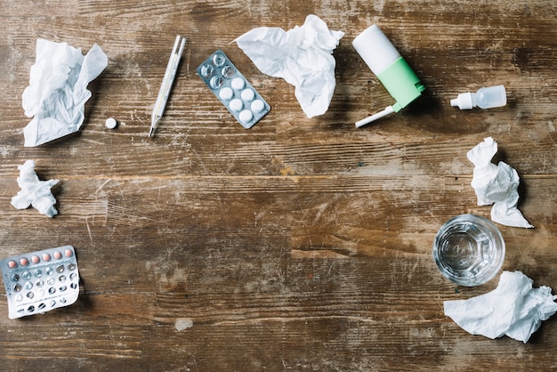 Elevated view of medicines; crumpled tissue paper; thermometer; throat spray and glass of water on wooden backdrop