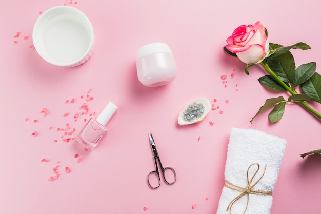 Elevated view of nail varnish; scissors; salt; towel; flowers and moisturizing cream on pink surface