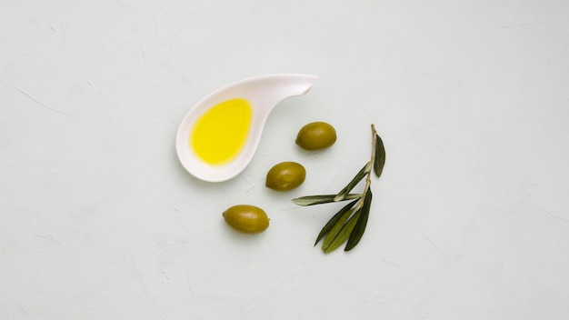 An elevated view of olive oil in white bowl with olives and leaves on concrete backdrop