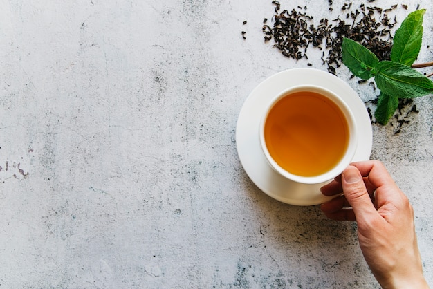 An elevated view of a person holding cup of tea with dried tea leaves and mint