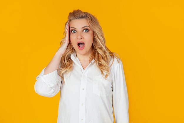 Embarrassed European woman blonde frowning rounds her eyes, holding her hand on her head with her mouth wide open