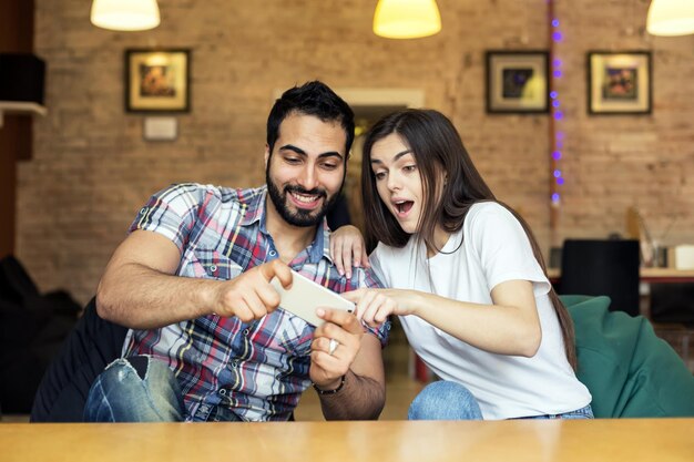 Emotional bearded handsome man and brunette girl having fun playing online game on smartphone in stylish room