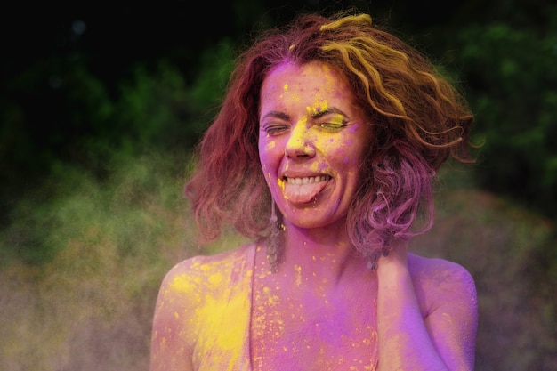 Emotional caucasian model with curly hair posing in a cloud of dry paint and showing her tongue