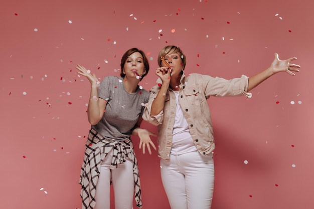 Photo emotional cool two women with short hair in white modern pants posing with confetti and looking into camera on isolated backdrop